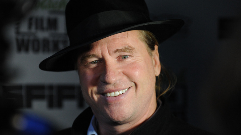 Val Kilmer smiling in a black hat with a dark background at an event