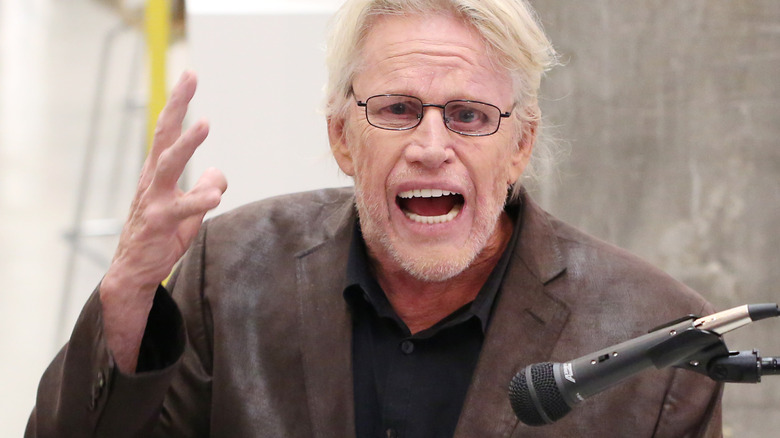 Gary Busey speaks emphatically in to a microphone and gestures with his hand