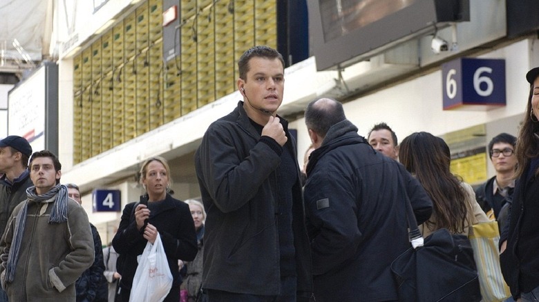 Jason Bourne speaks into an earpiece in a crowded station