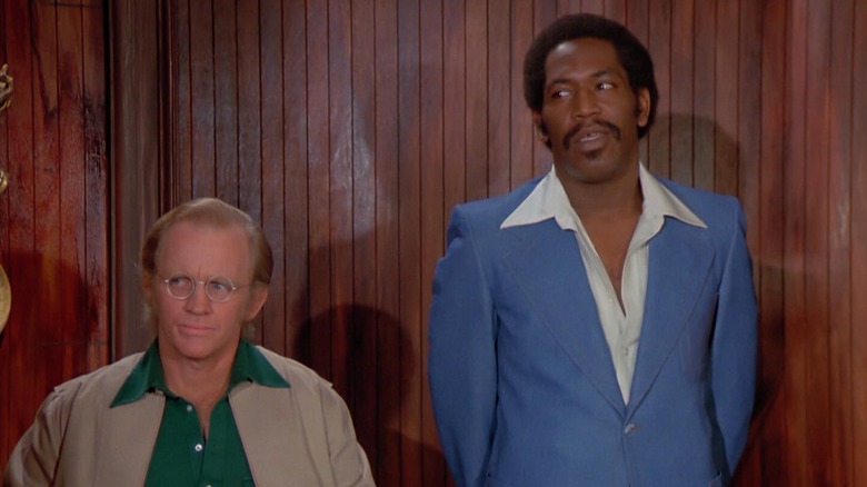 Bubba Smith and short man stand against wood paneled wall