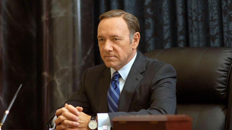 Frank Underwood sits at an ornate desk with his hands clasped