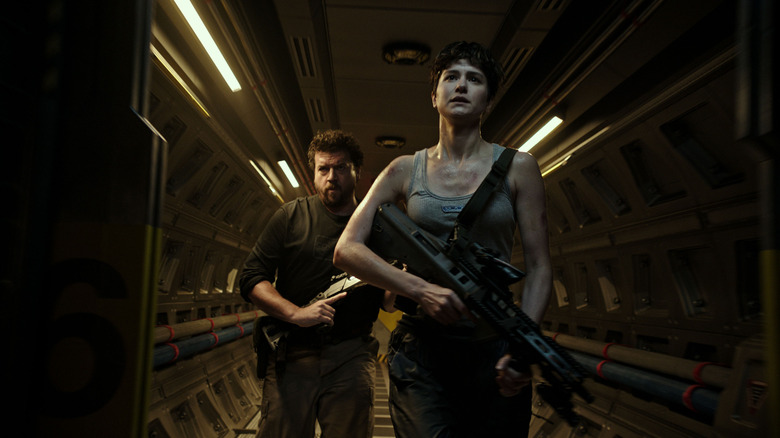 Danny McBride and Katherine Waterston walking down hallway with guns 