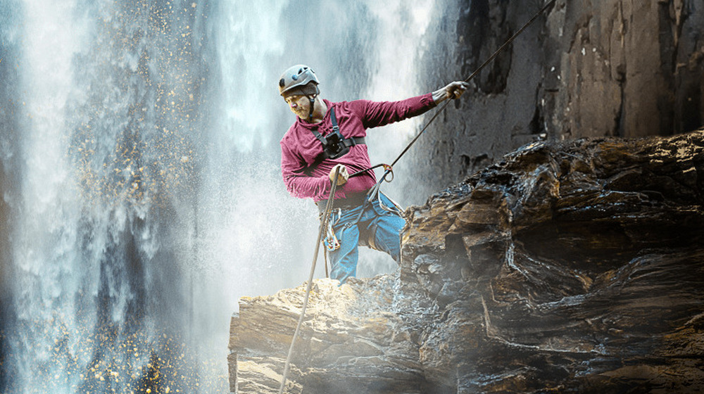 A miner on a guideline heads down the side of a cliff with a waterfall raging in the background.  Bright sunlight shines off of his body.