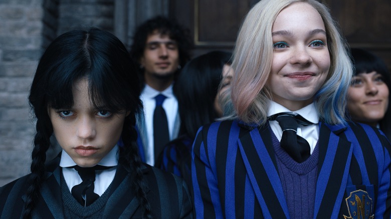 Enid smiling and Wednesday scowling as they stand in their school uniforms