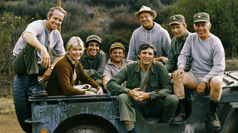 The original cast of MASH, smiling in a Jeep