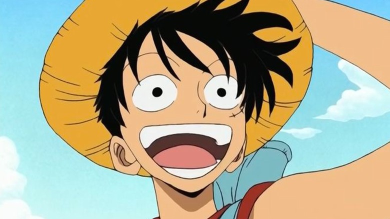 Luffy smilng while holding his straw hat under a blue sky with fluffy white clouds